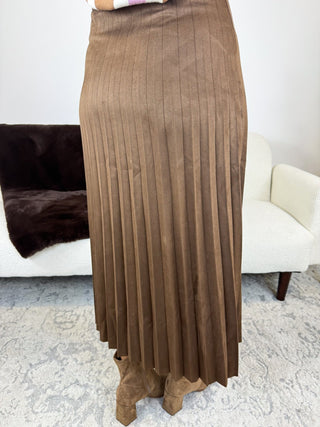 A brown pleated midi skirt shown on a model in a living room setting, paired with brown suede boots.