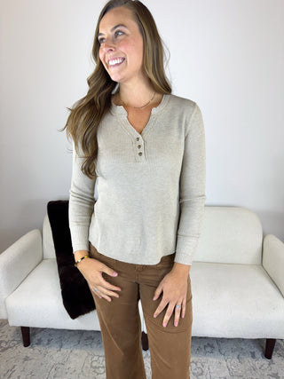 A woman modeling a light beige waffle-knit henley shirt with brown pants, standing in front of a white sofa.