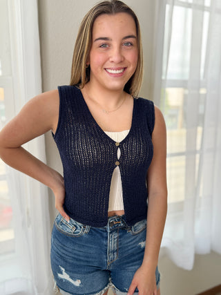 Woman wearing a navy blue knitted vest over a white top with denim shorts indoors.