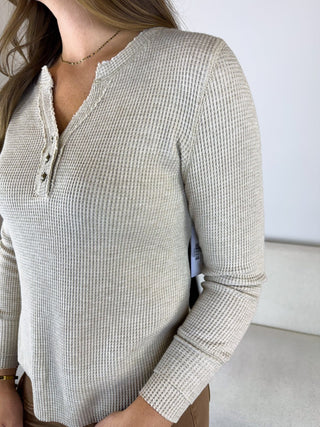 A woman wearing a light beige waffle-knit long-sleeve henley top with a buttoned split neckline, shown in a close-up indoor shot.