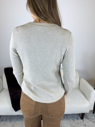 A woman viewed from behind wearing a light beige textured knit sweater and brown pants, standing in front of a white armchair in a bright indoor setting.