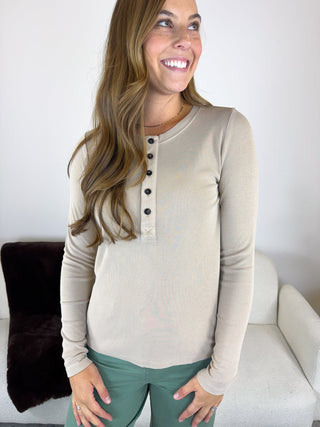A woman wearing a beige long-sleeve henley top with black buttons and sage green pants, standing in a brightly lit indoor setting.
