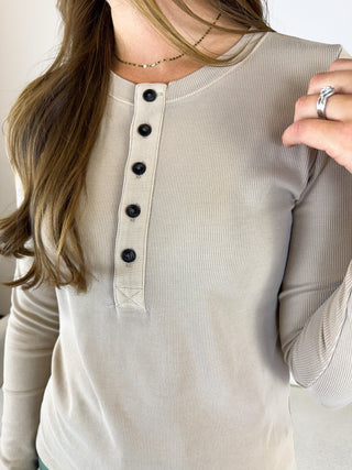 A woman wearing a beige ribbed long-sleeve henley top with a button placket and black buttons, shown in a close-up lifestyle shot.