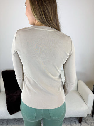 A woman viewed from behind wearing a fitted, long-sleeve ribbed knit top in a light cream or off-white color, paired with sage green pants, standing in a living room setting.