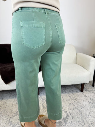 A person shown from the back wearing mint green cropped pants with a relaxed straight-leg fit, styled with a cream top and beige sandals in a living room setting.