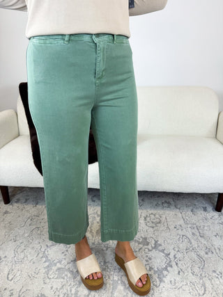 A person wearing sage green cropped wide-leg pants, paired with a cream top and cream slide sandals, standing in front of a white textured sofa on a patterned rug.