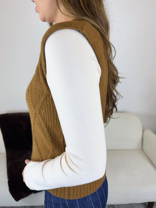 A woman shown from the side wearing a brown ribbed knit sleeveless vest over a white long-sleeve top, standing indoors near a light-colored sofa.