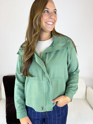 A woman wearing a sage green cropped utility jacket over a white t-shirt and blue plaid pants, standing in a brightly lit indoor setting.