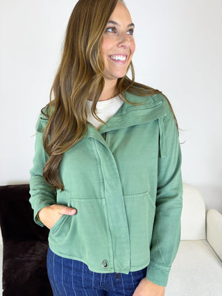 A woman wearing a sage green cropped jacket with a wide collar and front button closure, paired with blue jeans, standing in a brightly lit indoor setting.