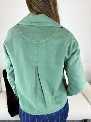 A woman viewed from behind wearing a sage green long-sleeve button-up shirt and blue pinstriped pants, standing indoors against a white wall.