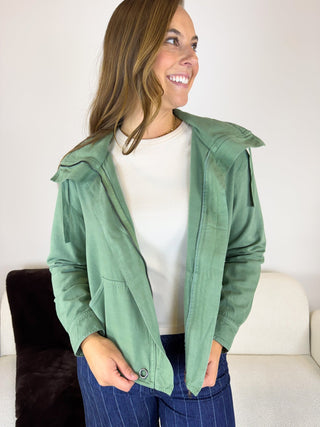 A woman wearing a sage green hooded jacket over a white t-shirt and blue pinstripe pants, standing in a brightly lit indoor setting.