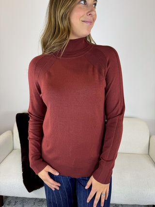 A woman wearing a ribbed mock neck long-sleeve sweater in a dusty terracotta red color, paired with dark blue pinstriped jeans, standing in front of a white couch.