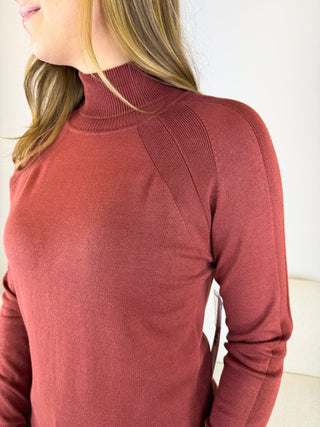 A close-up of a woman wearing a ribbed mock neck long-sleeve sweater in a dusty rose or terracotta color, showing the texture and fit against a plain light background.