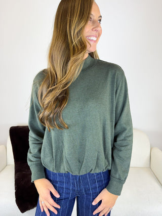 A woman wearing a sage green mock neck long-sleeve top and blue pinstripe jeans, standing in a brightly lit room with a white sofa and a dark brown pillow.