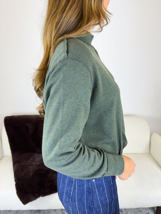 A woman shown from the side wearing a sage green mock neck long-sleeve top and blue pinstriped pants, standing in front of a white sofa with a dark brown throw pillow.