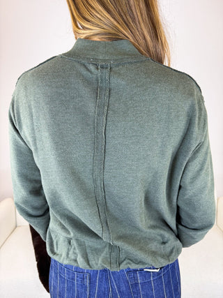A person viewed from the back wearing a muted sage green knit top or sweater with a high mock neck and a central seam running down the back, paired with blue pinstriped pants.
