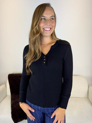 A smiling woman with long light brown hair stands indoors wearing a black ribbed long-sleeve henley top with a three-button placket and blue pinstripe jeans.