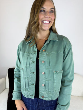 A woman smiling while wearing a sage green cropped denim jacket over a black top and blue jeans, standing indoors against a white wall.
