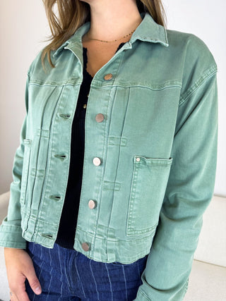A woman wearing a cropped sage green denim jacket over a black top and blue jeans, shown from the neck down in a clean indoor setting.