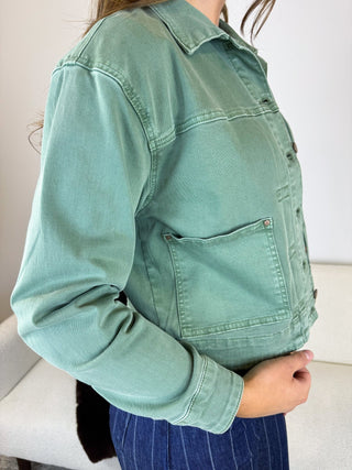 A person wearing a sage green cropped denim-style jacket with a button front and chest pocket, paired with blue pinstriped pants, photographed indoors against a light wall and sofa.