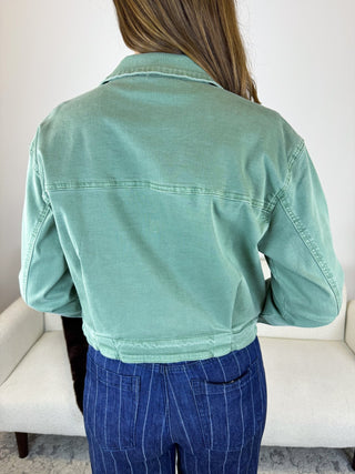 A person shown from the back wearing a cropped sage green denim jacket and blue pinstripe pants, standing in front of a white upholstered chair.