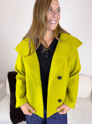 A woman wearing a bright lime green oversized coat over a black top and blue pinstripe pants, standing in front of a white sofa.