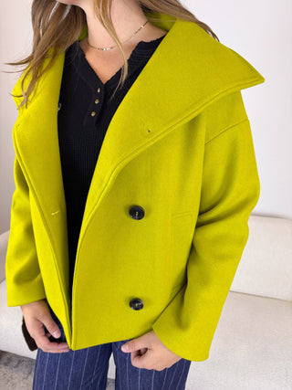 A woman wearing a bright yellow-green oversized coat over a black henley top and blue pinstripe pants, shown from the neck down in an indoor setting.