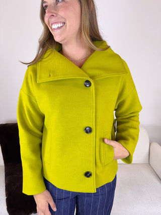 A woman wearing a bright lime green wool-blend coat with a wide collar and three dark buttons, paired with blue pinstripe pants, standing indoors against a white wall.