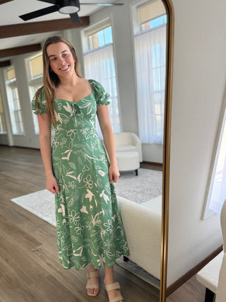 Woman in a green floral dress standing in a room with a mirror.