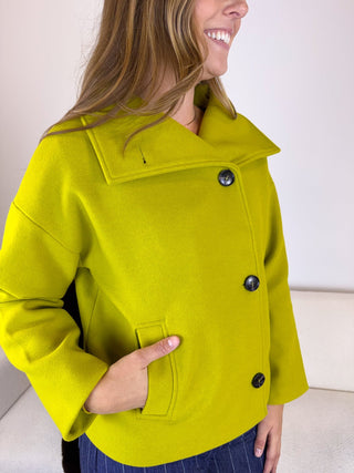A woman wearing a bright lime green wool-blend coat with a wide collar and dark buttons, standing indoors against a light neutral wall.