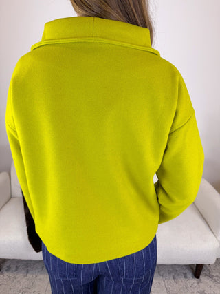 A person shown from the back wearing a bright lime green long-sleeve top or lightweight jacket, paired with blue pinstriped pants, standing indoors in front of a white sofa.