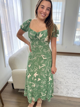 Woman wearing a green floral dress in a room with white curtains and a beige chair.