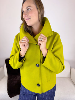 A woman wearing a bright lime green wool-blend coat with a large collar and dark buttons, standing in front of a white sofa.