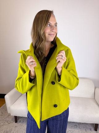 A woman wearing a bright lime green double-breasted coat over a black top and blue pinstripe pants, standing in front of a white sofa.
