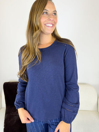 A woman wearing a navy blue long-sleeve top with eyelet lace detailing on the cuffs and shoulders, paired with blue jeans, standing in a brightly lit indoor setting.