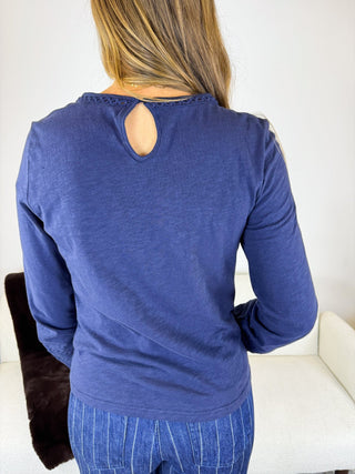 A woman viewed from behind wearing a blue long-sleeve top with a keyhole cutout at the back and blue pinstriped pants, standing indoors against a light wall.