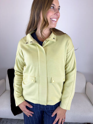 A woman wearing a light yellow-green cropped jacket with a stand collar and button details, paired with dark blue jeans, standing in front of a light-colored sofa.