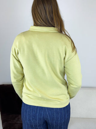 A person shown from the back wearing a pale yellow-green long-sleeve top and blue pinstriped pants, standing indoors against a light wall.