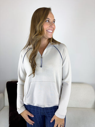 A woman wearing a cream-colored quarter-zip sweatshirt with dark blue contrast piping and a matching zipper pull, paired with blue jeans, standing in front of a light-colored wall and sofa.