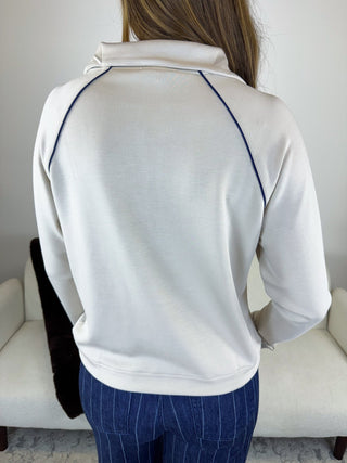 A person viewed from behind wearing a white long-sleeve quarter-zip pullover with dark blue contrast stitching along the shoulders and sleeves, paired with blue pinstriped pants, standing in front of a light-colored armchair.