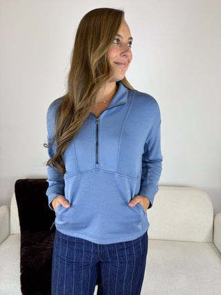 A woman wearing a dusty blue quarter-zip pullover with a front kangaroo pocket, paired with dark blue pinstripe pants, standing in front of a white couch.