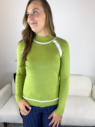 A woman wearing a lime green mock neck knit sweater with white contrast trim and raglan sleeves, paired with dark blue pinstriped pants, standing in front of a white couch.