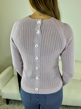 A woman viewed from the back wearing a light lavender open-knit cardigan with a vertical row of white buttons down the center, paired with blue pinstriped pants in an indoor setting.