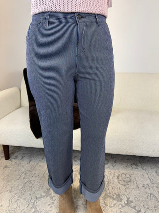 A person wearing high-waisted, straight-leg blue jeans with thin white vertical pinstripes and rolled cuffs, paired with a pink knit top and tan boots, standing in front of a white couch.