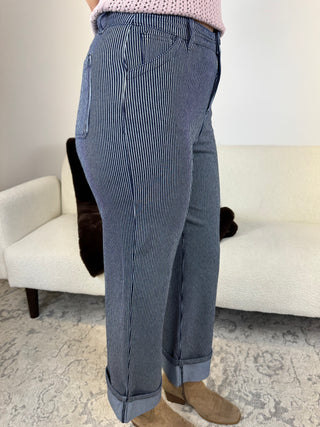 A person wearing blue-gray pinstriped jeans with a pink knit top and tan boots, standing in a living room in front of a white textured sofa.