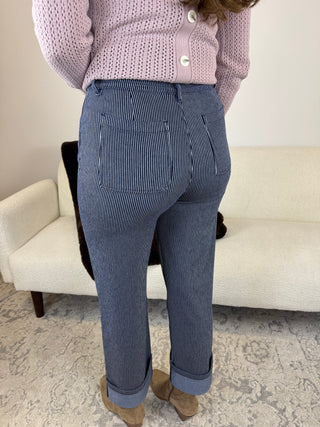 A person shown from the back wearing blue-and-white pinstriped trousers, a light pink knit cardigan, and tan ankle boots, standing in front of a cream-colored sofa.