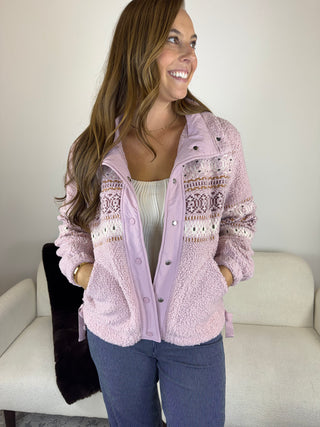 A woman wearing a light pink fuzzy button-up cardigan with a white and brown Fair Isle pattern on the chest, paired with a white top and blue jeans, standing in front of a light-colored sofa.