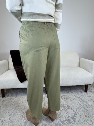 A person shown from the back wearing olive green wide-leg trousers, a cream and gray striped sweater, and tan boots, standing in a living room with a white sofa and patterned rug.