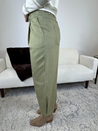 A person wearing olive green wide-leg trousers, styled with a cream knit sweater and brown suede boots, standing in a living room setting with a white textured sofa in the background.