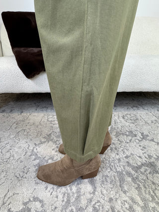 A person wearing light olive green trousers and taupe suede ankle boots, standing on a gray patterned rug in front of a white sofa with a dark brown throw.